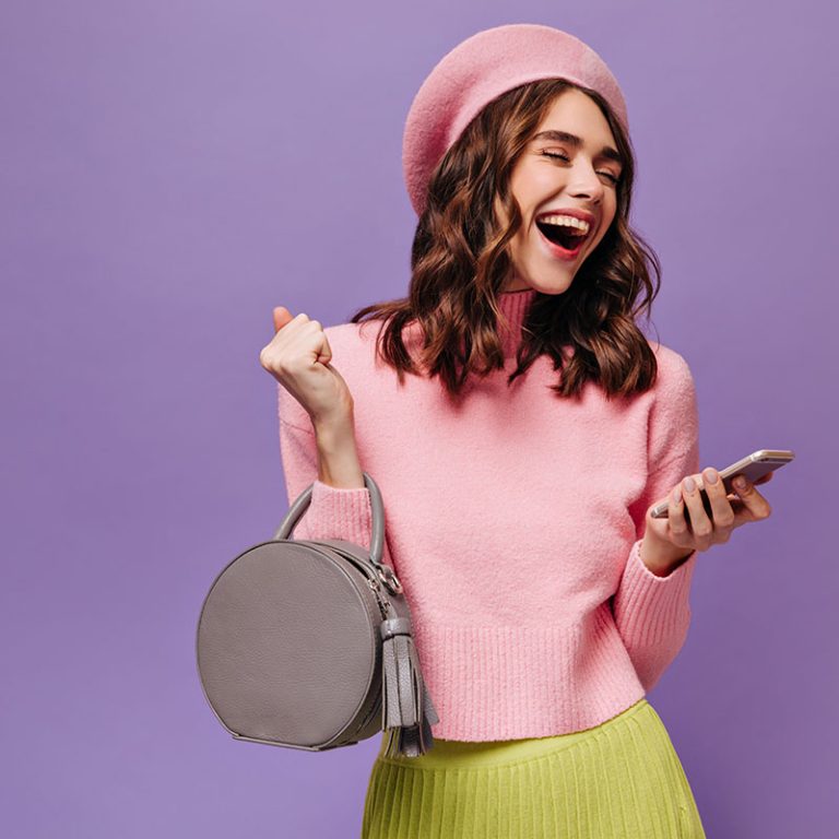 Happy lady in pink beret, sweater and green skirt smiles and reads messages on phone. Cheerful gl holds grey handbag on purple background.